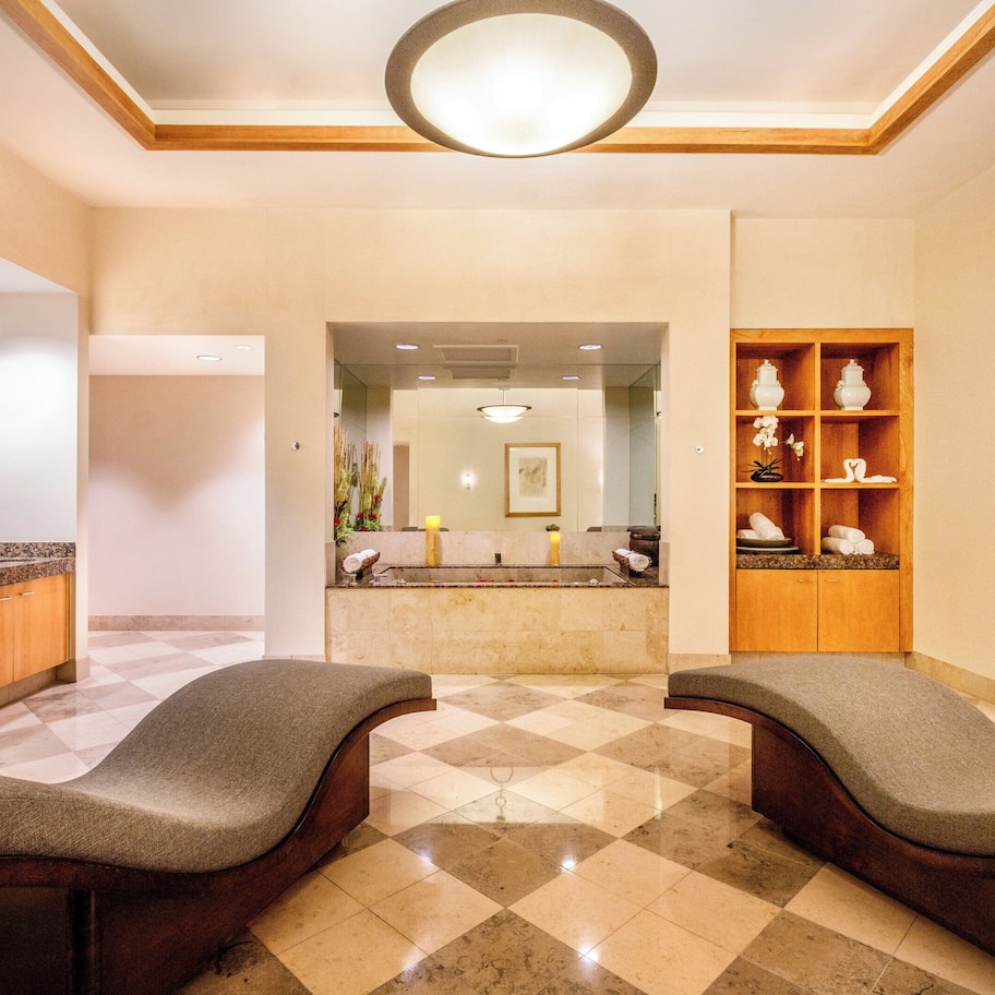 Un vestíbulo de hotel con piso a cuadros, dos zonas de estar con cojines, un mostrador de recepción y una estantería de madera con objetos decorativos.