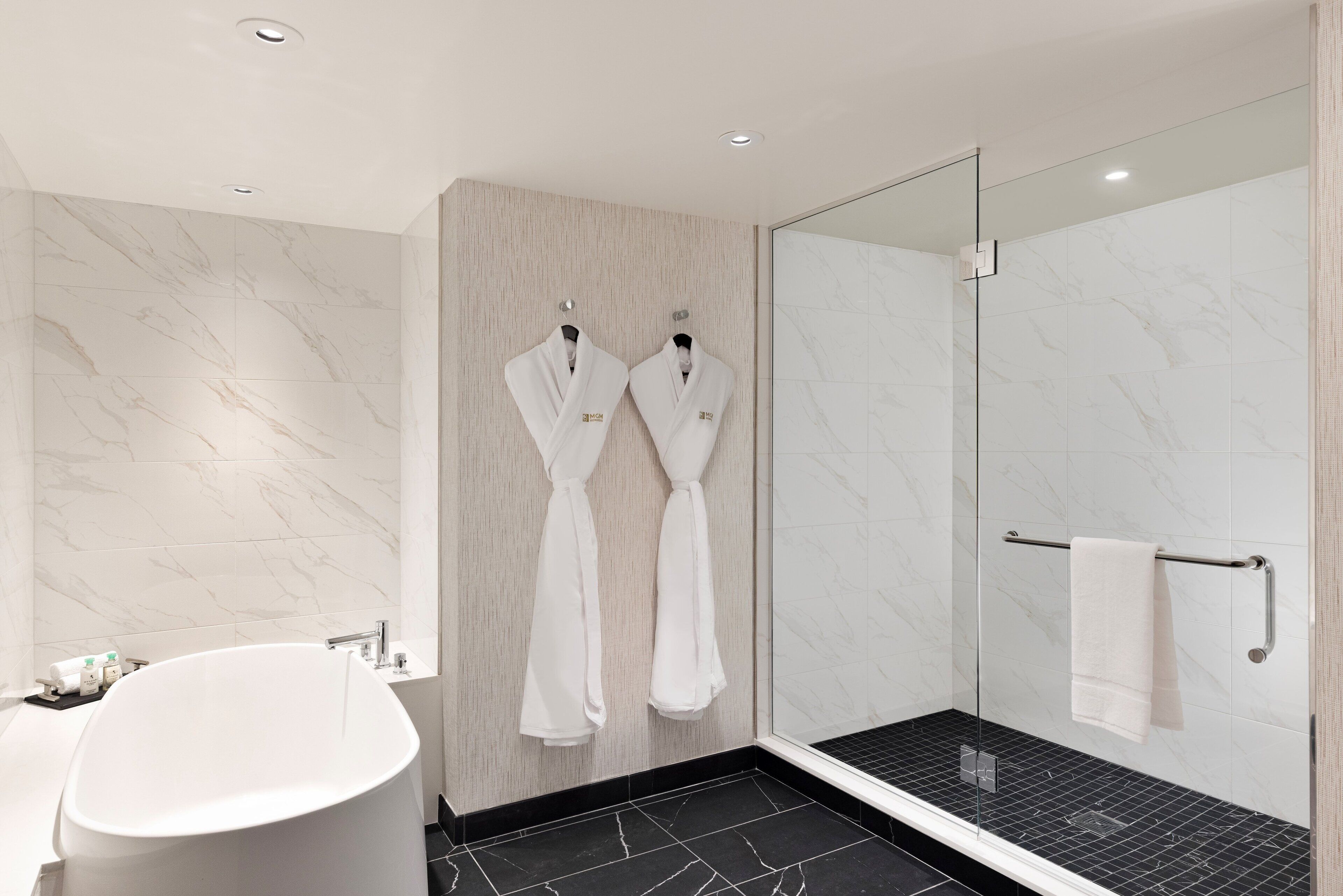 Un baño moderno con una bañera independiente, una ducha con cerramiento de vidrio y dos albornoces blancos colgados en ganchos.