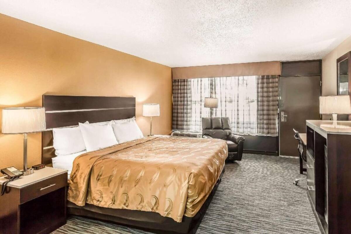 A hotel room with a bed, bedside lamps, a desk with a lamp, a chair, and a large window with curtains.