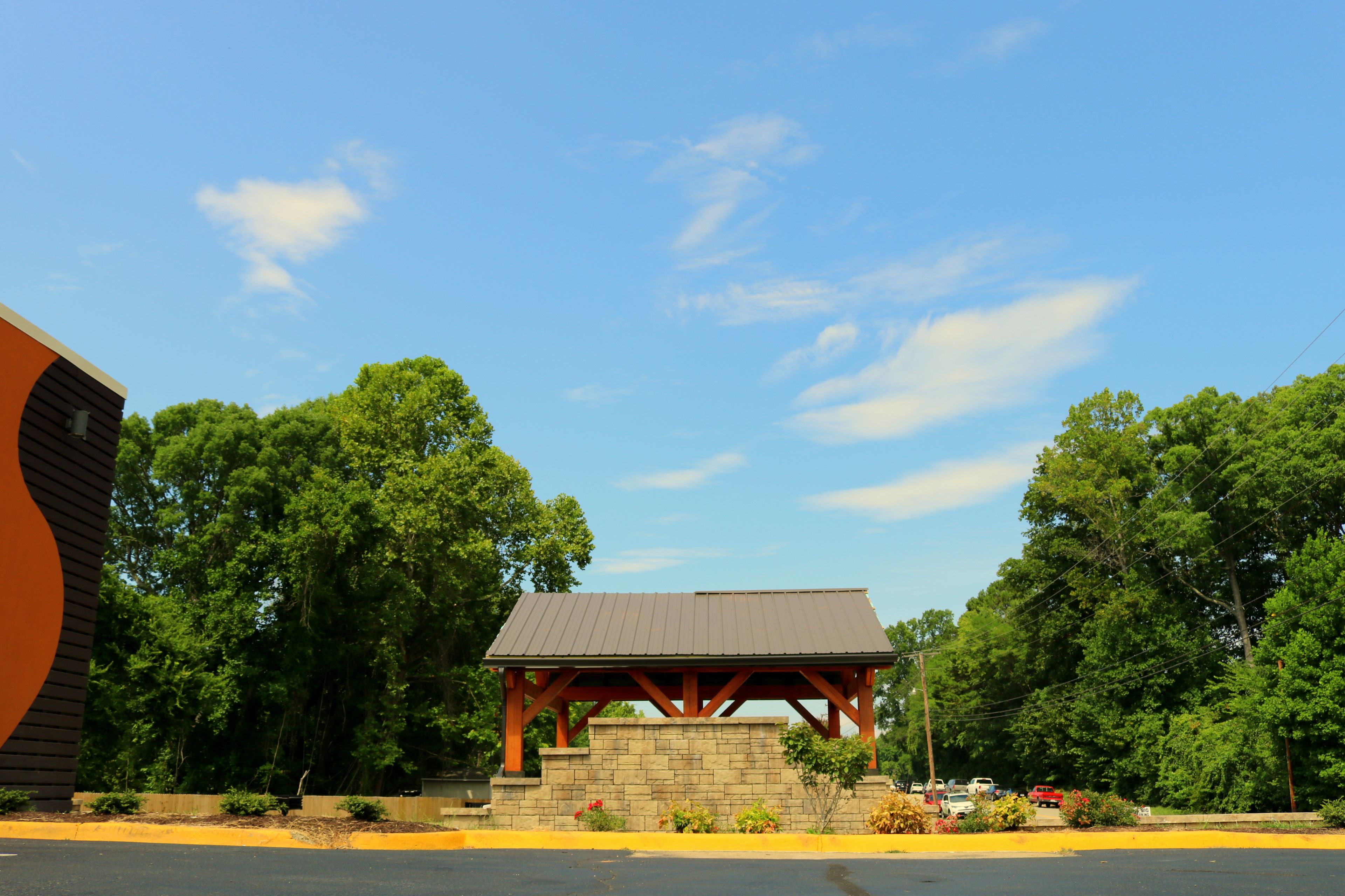 A pavilion with a sloped roof, surrounded by lush greenery and a clear blue sky.