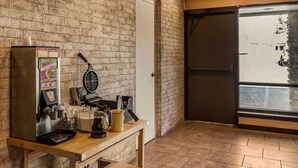Een bakstenen muur, een wafelijzer, een koffieapparaat en een raam met uitzicht op een straat.