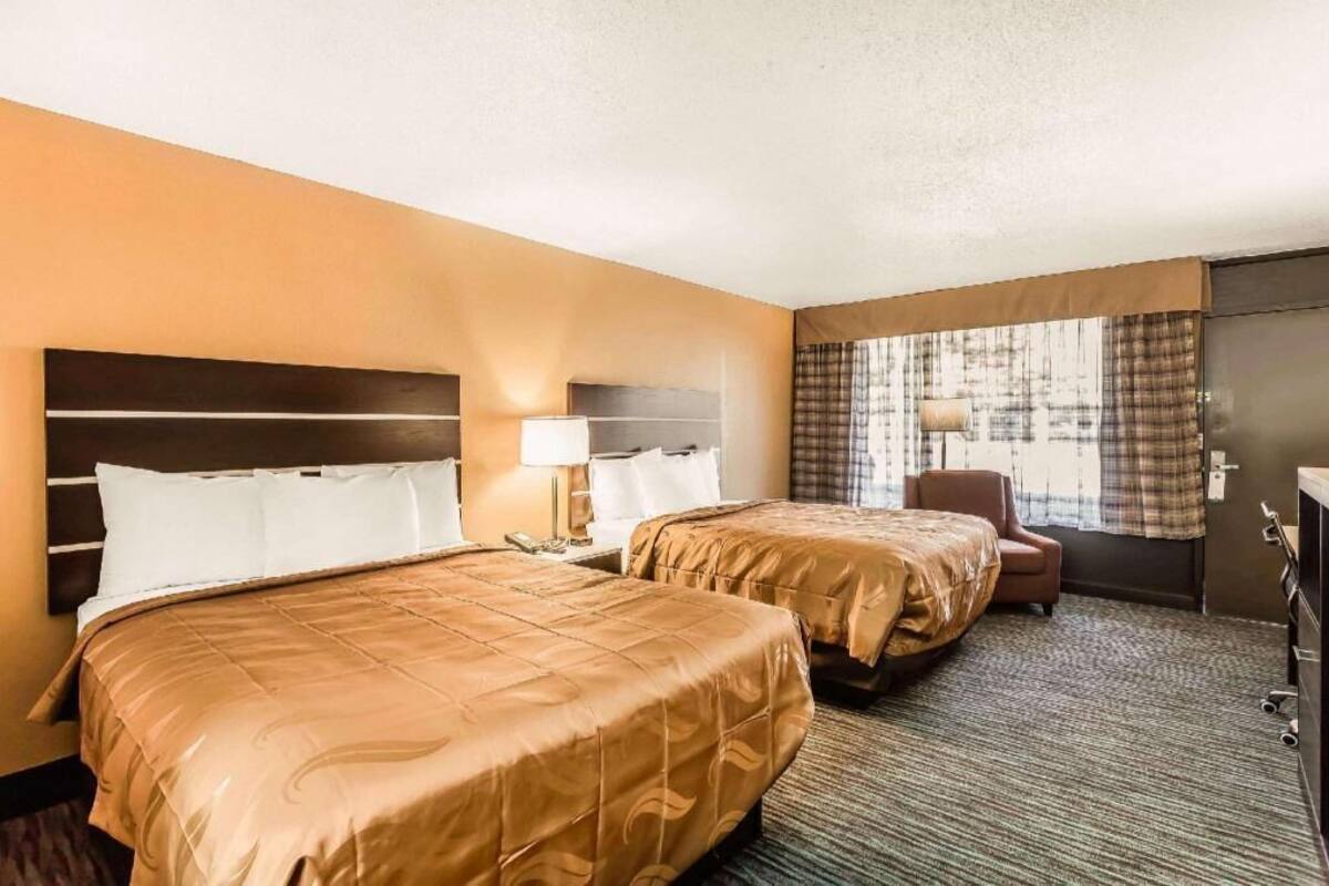A hotel room with two beds, a desk, a chair, a lamp, and a window with curtains.