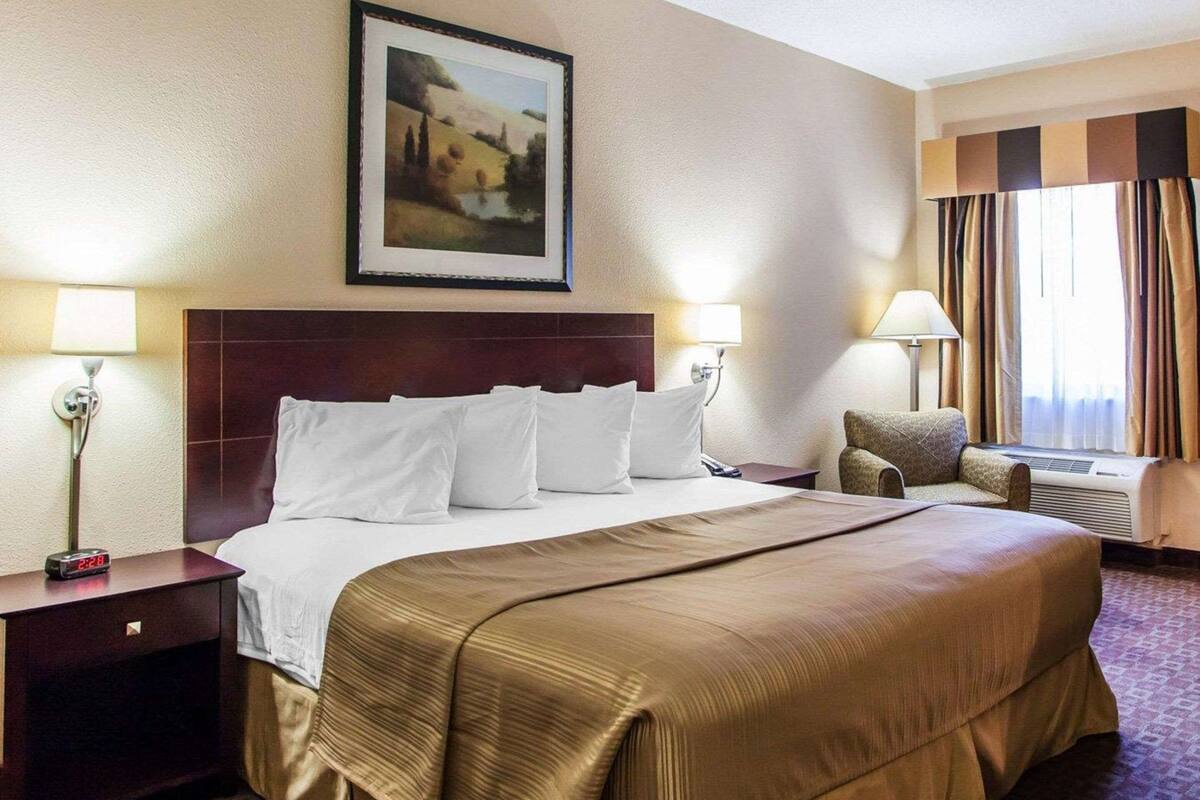 Habitación de hotel con una cama grande, dos mesitas de noche con lámparas, una silla, una ventana con cortinas y un cuadro en la pared.