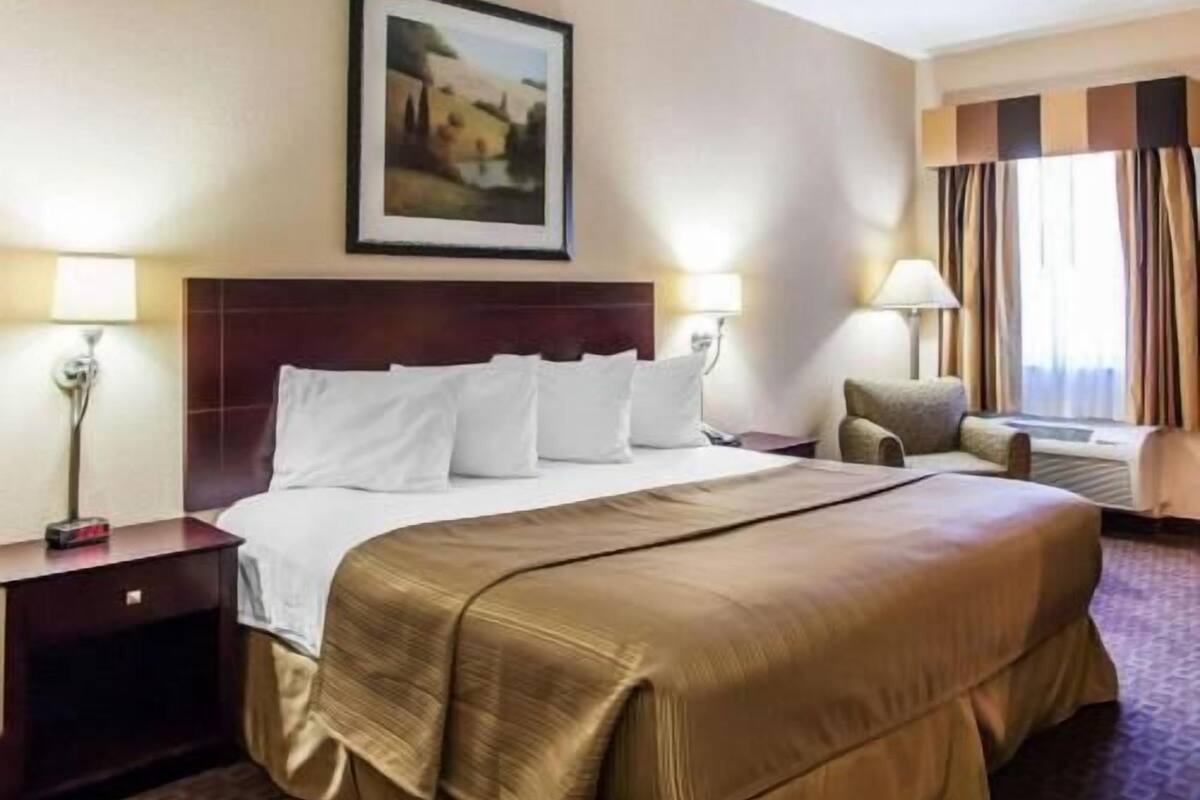 Habitación de hotel con una cama grande, dos lámparas de noche, una silla, un cuadro en la pared y una ventana con cortinas.