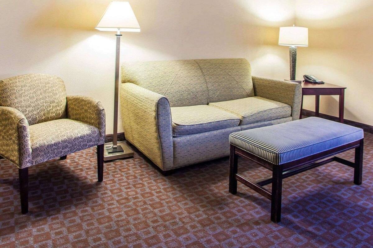 Una habitación de hotel con alfombra estampada, un sofá, una silla y dos mesitas de noche con lámparas.