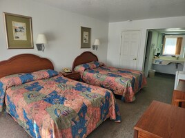 Habitación de hotel con dos camas, cabecero de madera, mesita de noche con lámpara, una pequeña mesa y un baño visible a través de una puerta abierta.