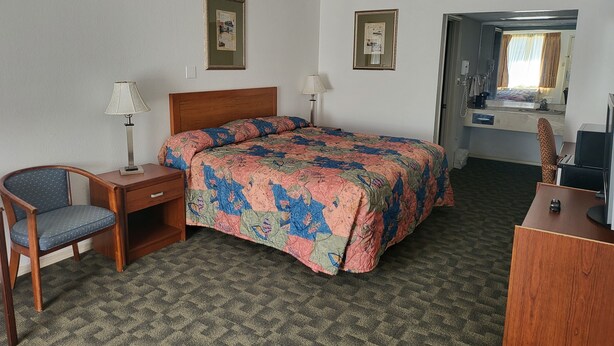 A hotel room with a bed, a desk, a chair, a desk lamp, a bedside table, and a mirror. - Blue Ribbon Inn and Suites (Sallisaw)