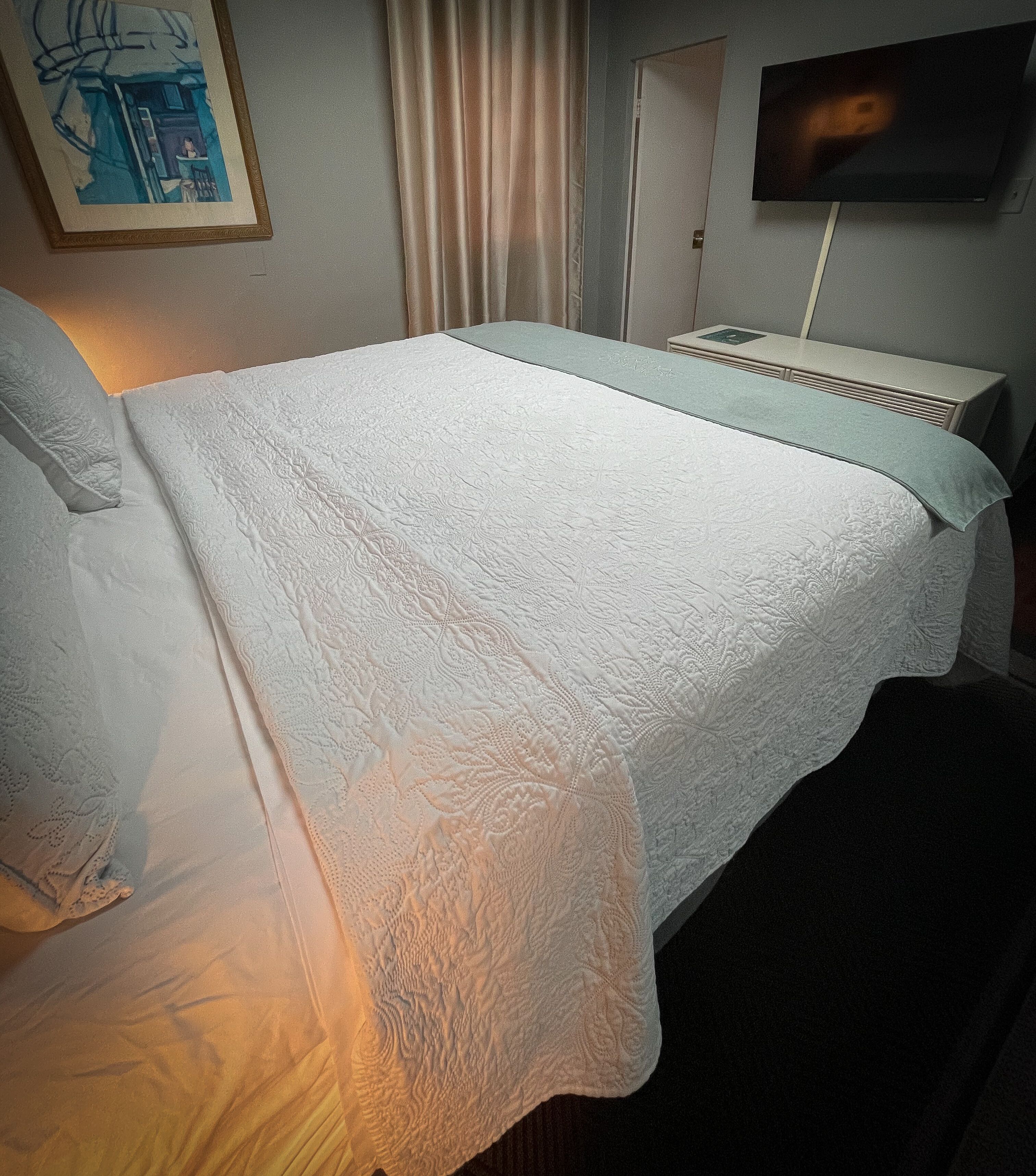 Una cama bien hecha con cubrecama y almohadas blancas, una mesita de noche con lámpara, un televisor en la pared y un cuadro en la pared.
