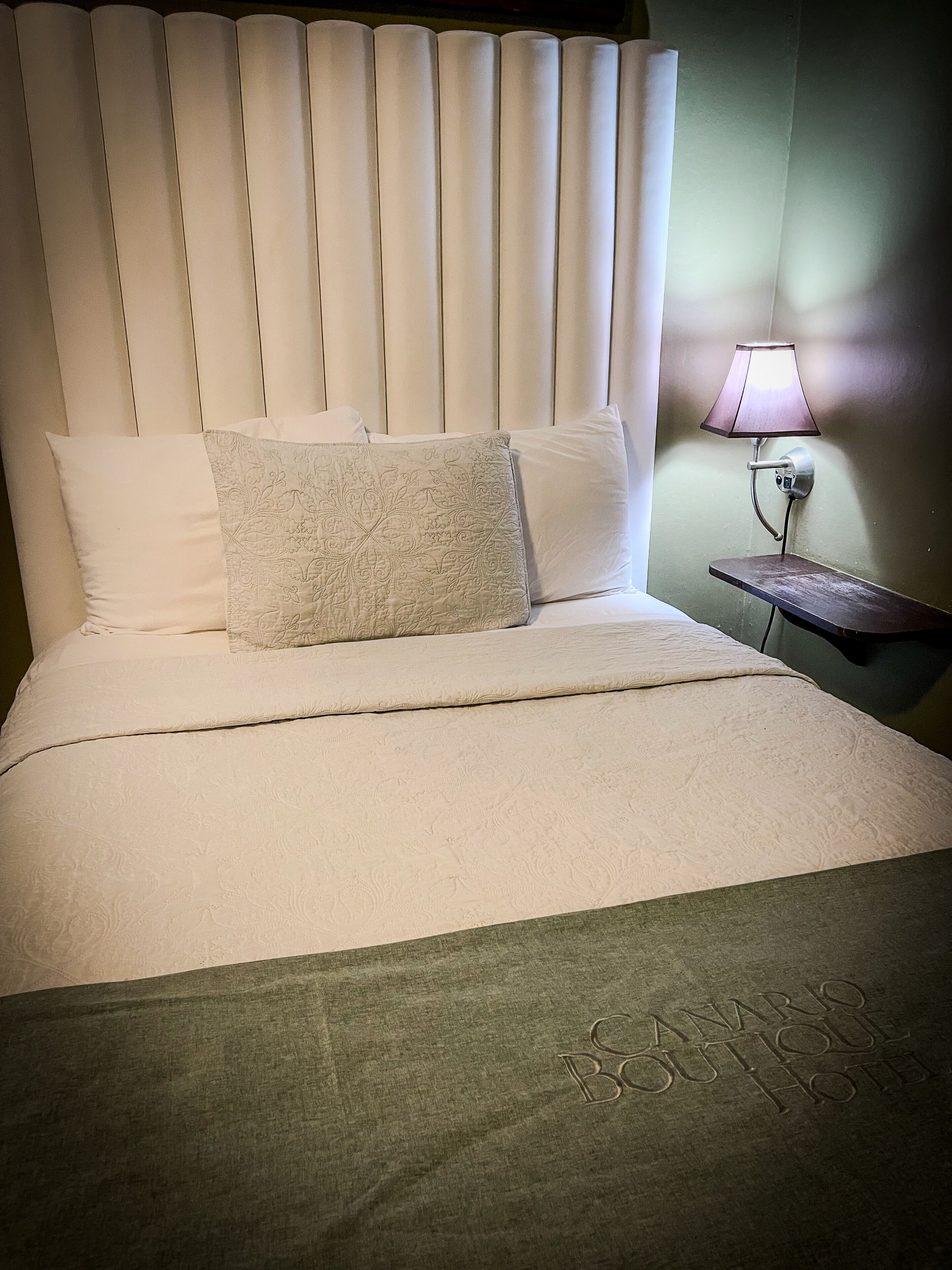 Een net opgemaakt bed met een gekleurd kussen, een hoofdbord en een bedlampje.