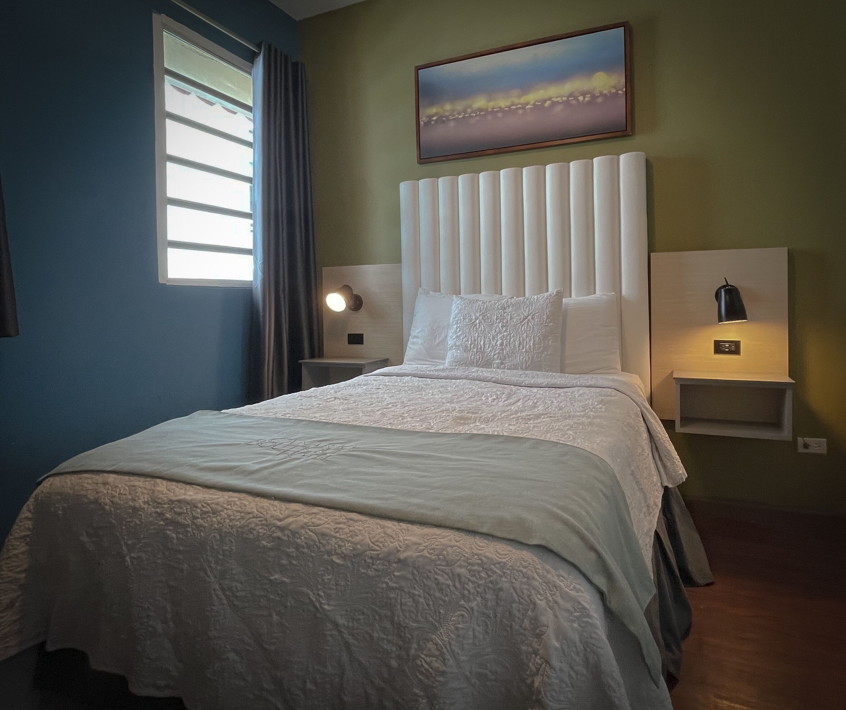 Una cama bien hecha con un cabecero blanco, dos mesitas de noche con lámparas, un cuadro en la pared y una ventana con cortinas.