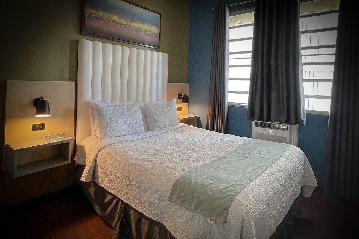Een net opgemaakt bed met een wit en lichtblauwe sprei, twee bedlampen, een nachtkastje en een schilderij aan de muur.