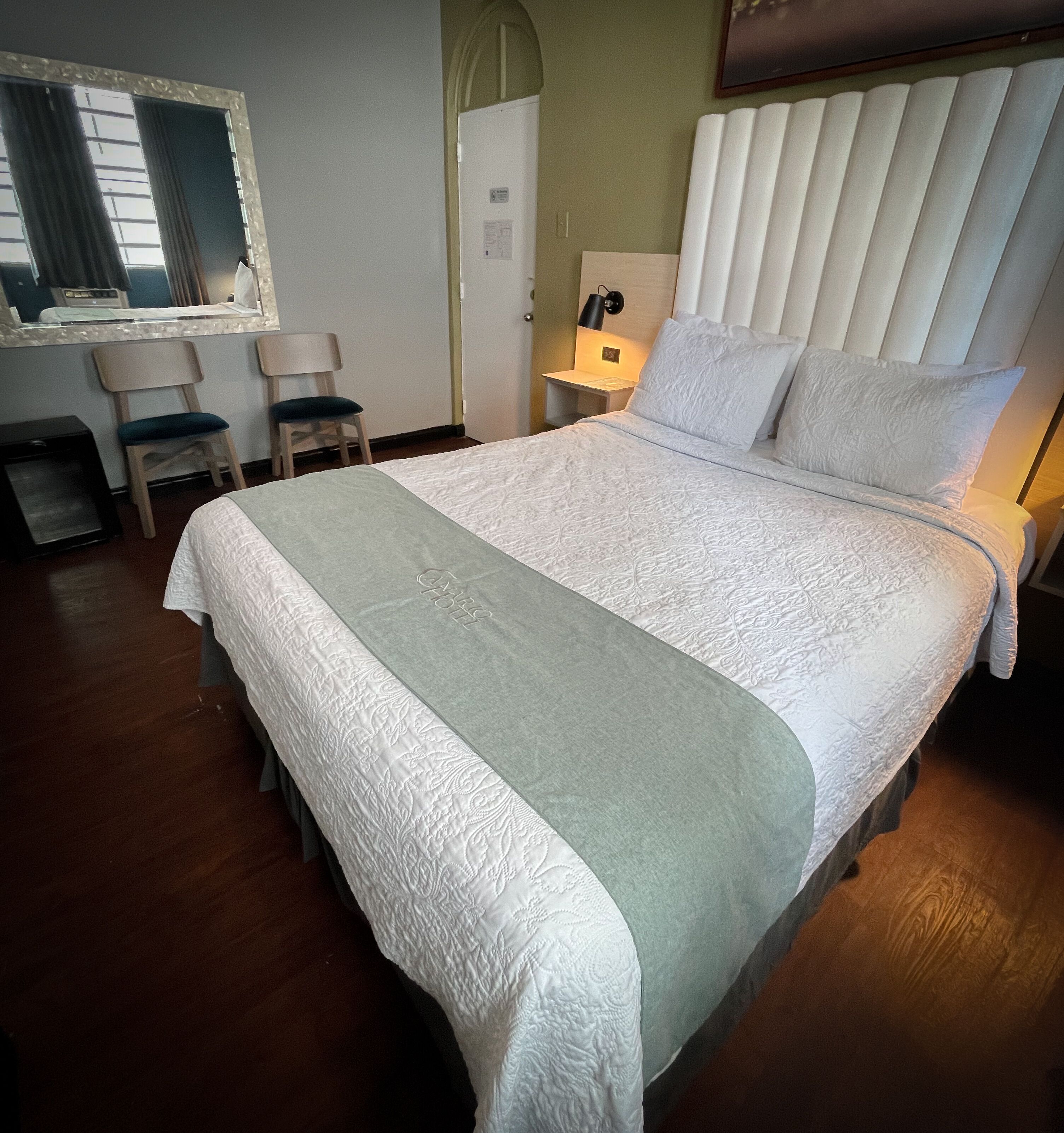 Un lit bien fait, recouvert d’une couvre-lit blanche et vert clair, avec une tête de lit en bois et une table de chevet sur laquelle se trouve une lampe. La pièce contient également deux chaises et une petite table avec un miroir.