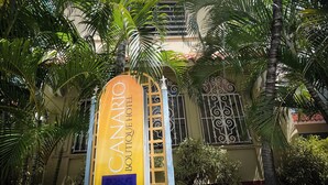 A hotel entrance with a prominent sign for the Canario Boutique Hotel, surrounded by lush palm trees and a traditional architectural style.