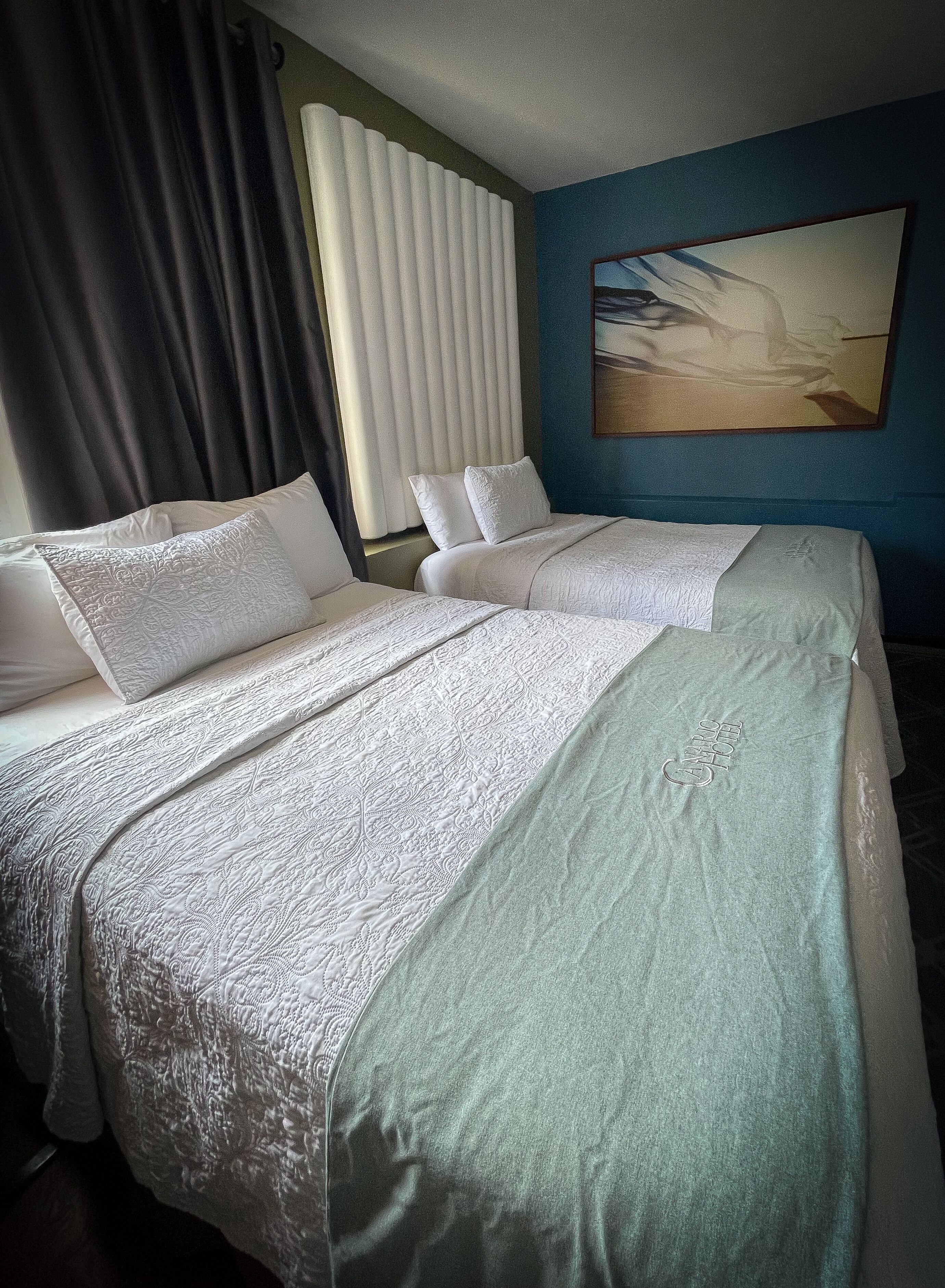 A hotel room with two beds, a large window with blinds, a painting on the wall, and a blue accent wall.