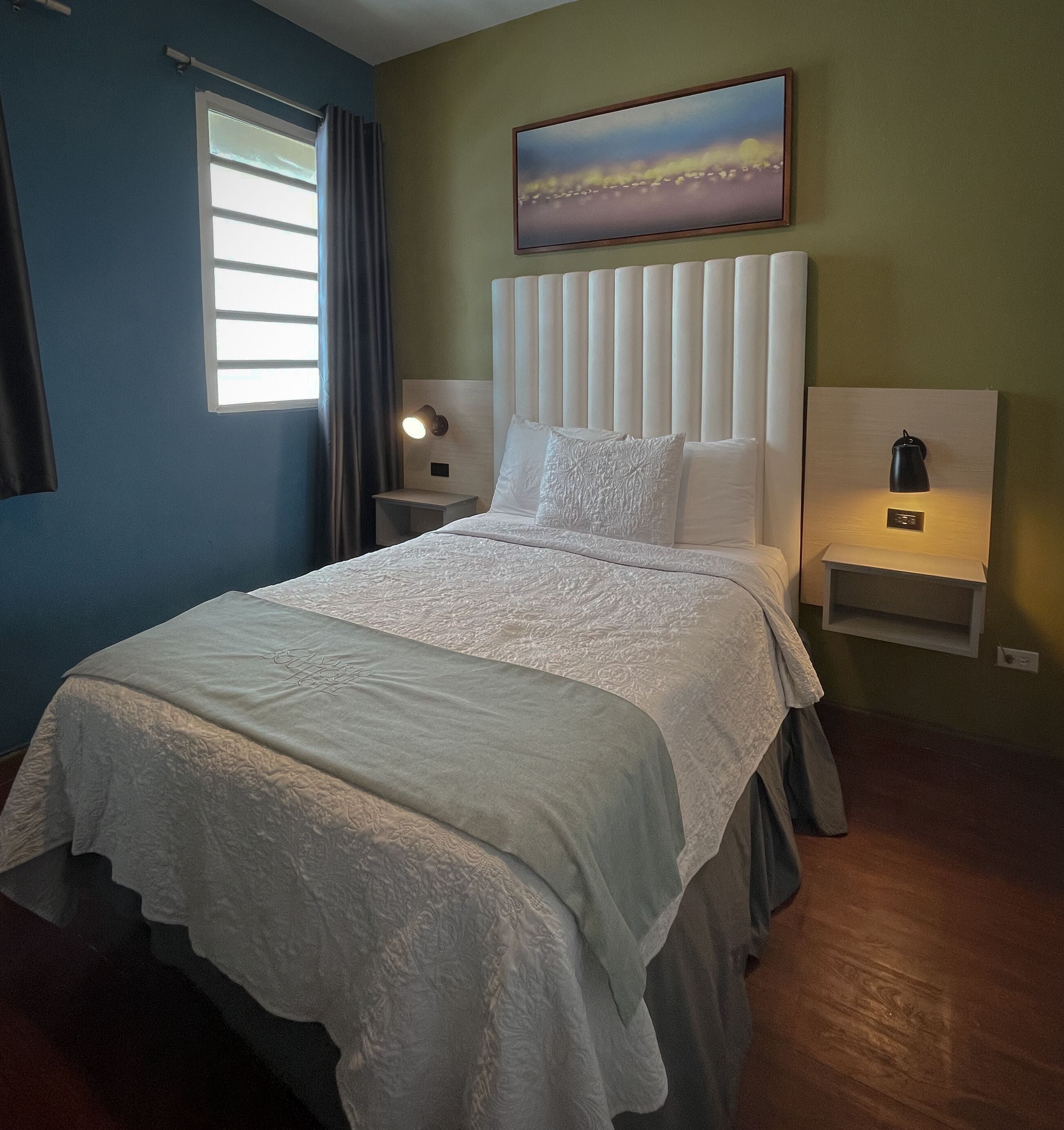 A neatly made bed with a white bedspread and pillows, a headboard, bedside tables with lamps, a window with curtains, and a painting on the wall.