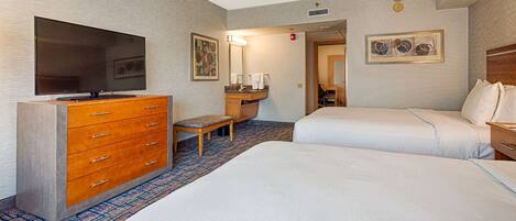 A hotel room with two beds, a TV, a wooden dresser, a bench, a bathroom with a sink and mirror, and artwork on the walls.