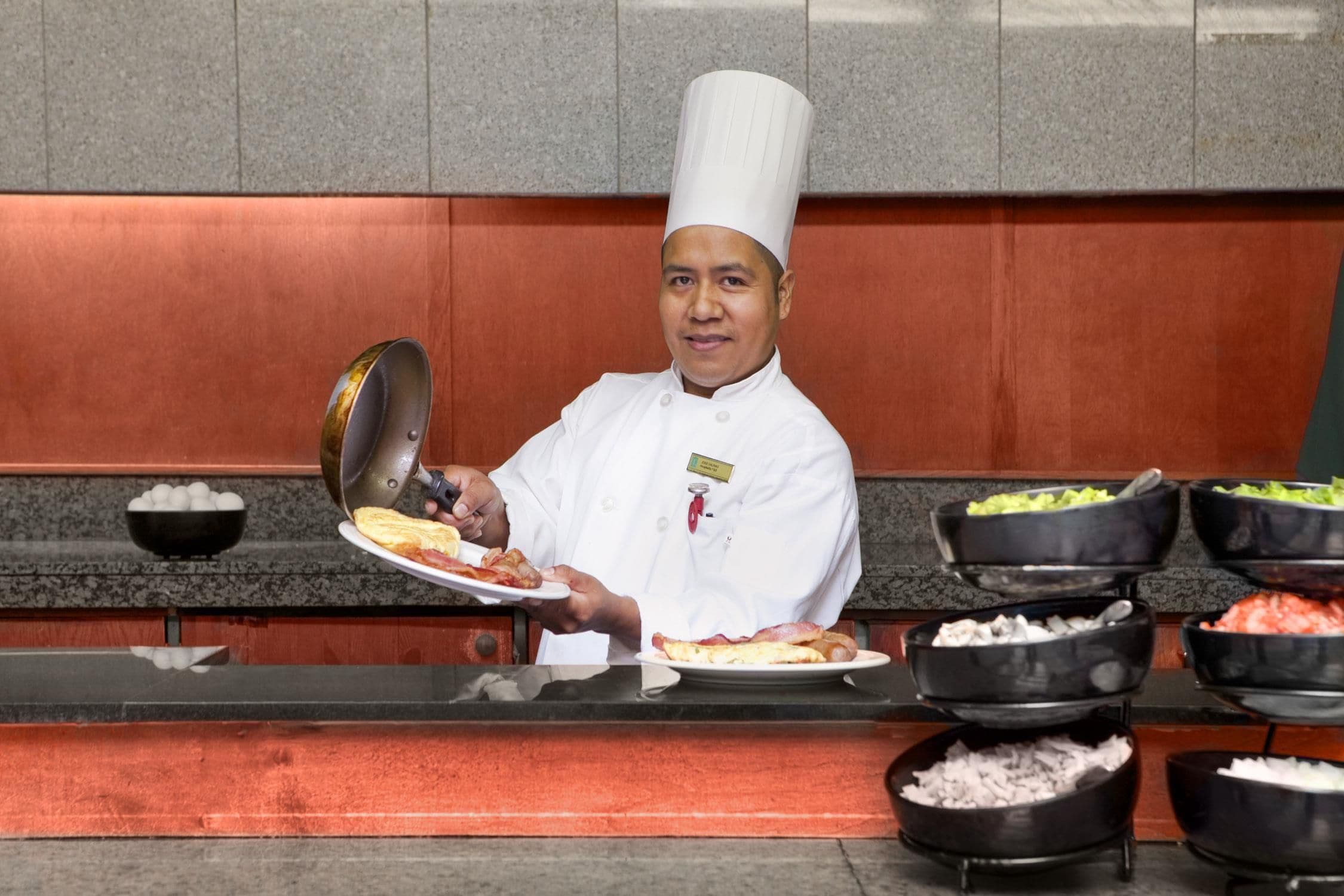 A chef in a white uniform holding a plate with food and a pan.