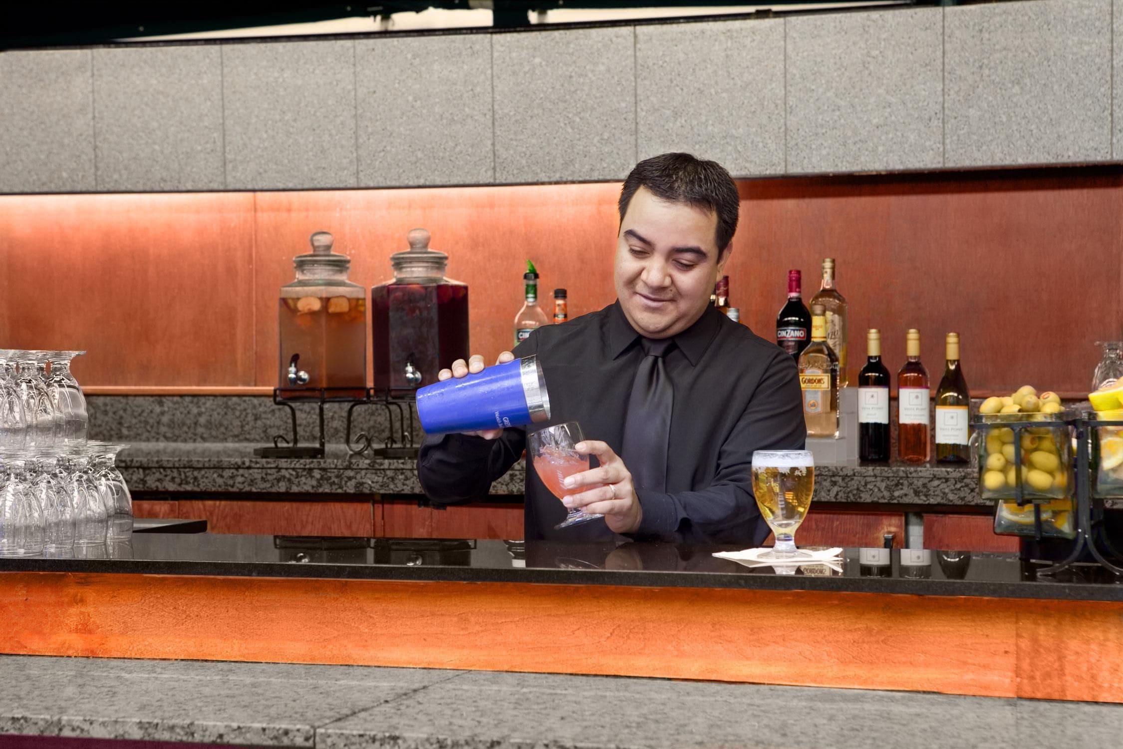 Un cantinero preparando una bebida detrás de la barra, con varias botellas y una presentación de frutas.