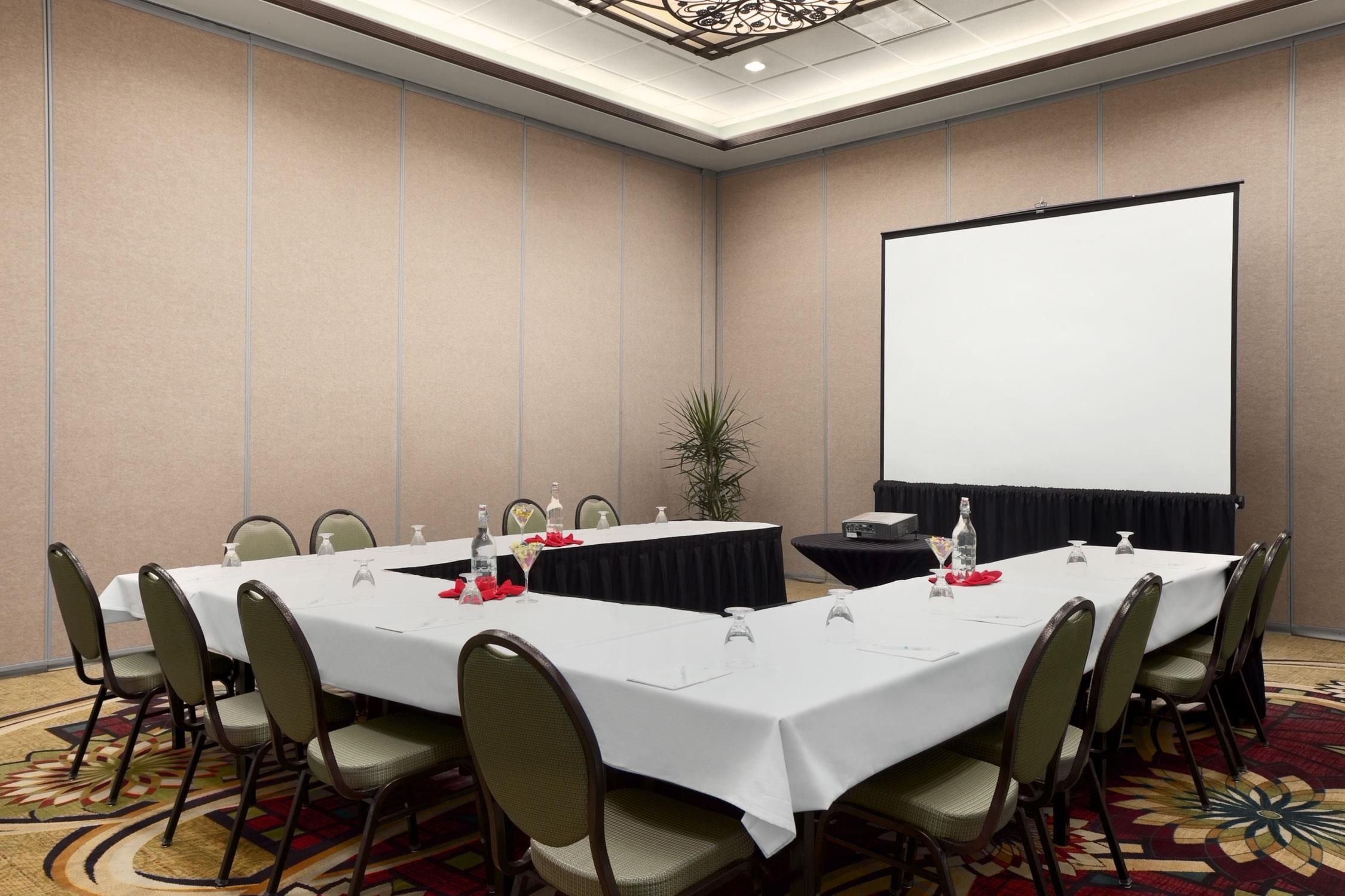 A conference room with a large table, chairs, a projector screen, and a plant.