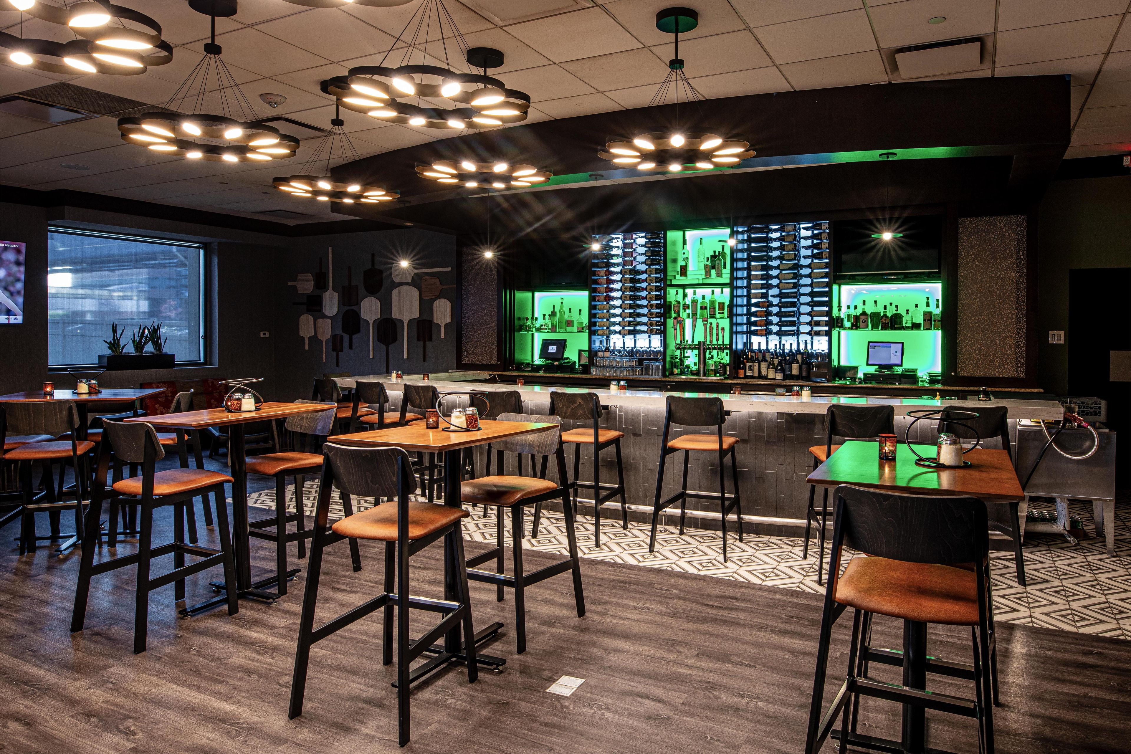 Zona de bar con diversas opciones de asientos, barra con iluminación verde y una pantalla de proyección en la pared.