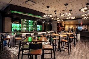 Zona bar moderna con bancone illuminato di verde, diverse lampade a sospensione e varie opzioni di posti a sedere, tra cui tavoli e sedie.