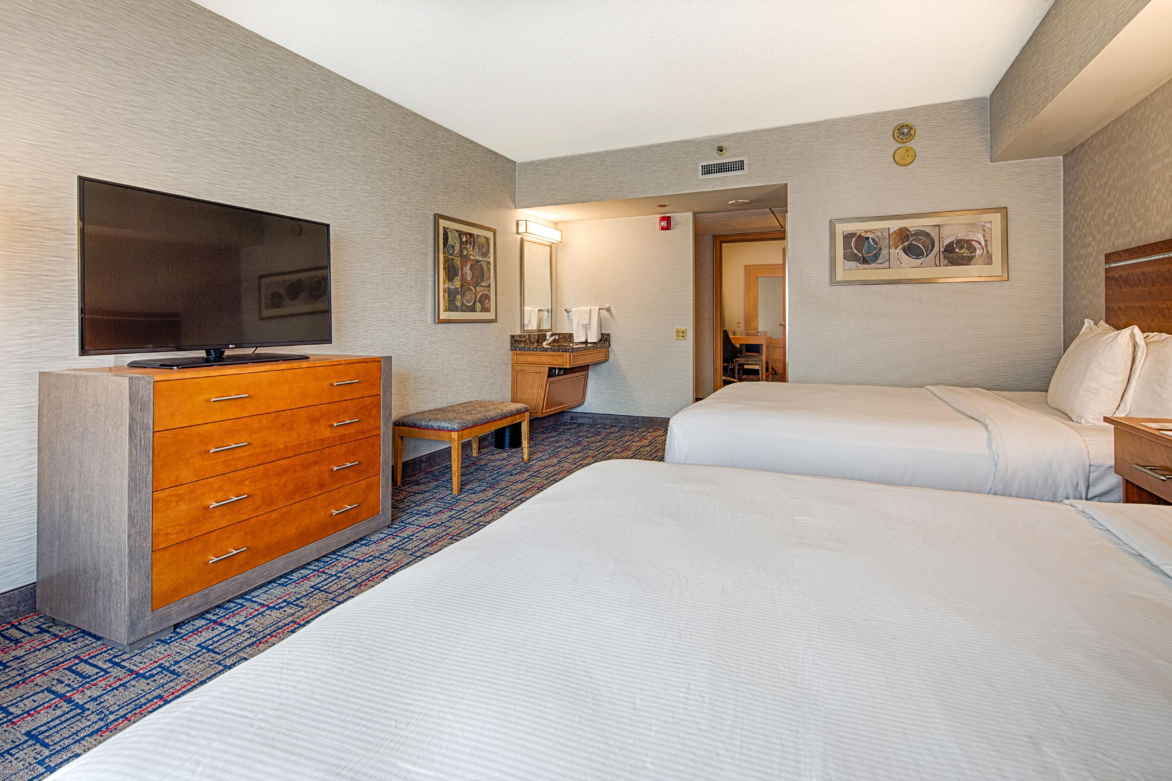 Habitación de hotel con televisor de pantalla plana, cómoda de madera, banca, baño con lavamanos y espejo, y dos camas.