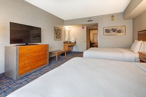 Habitación de hotel con televisor de pantalla plana, cómoda de madera, banca, baño con lavamanos y espejo, y dos camas.