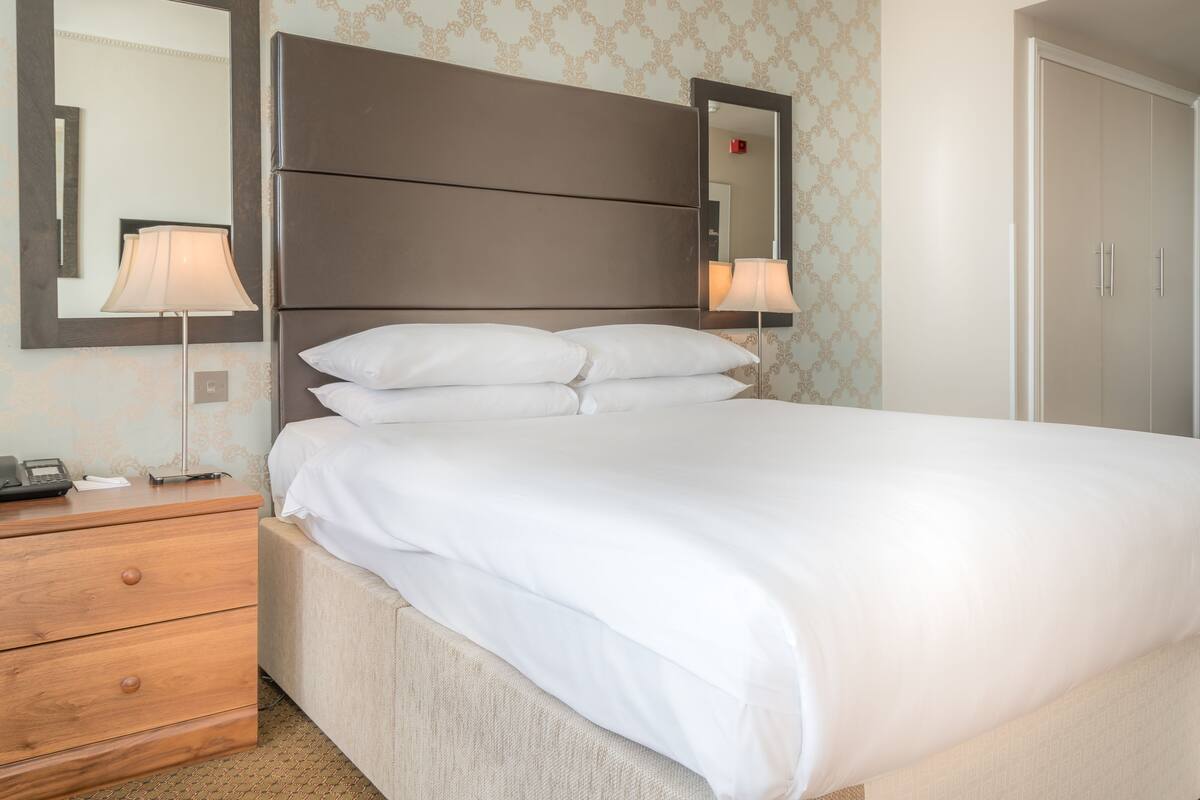 A neatly made bed with white linens, a wooden nightstand with a lamp, and a large mirror above the bed.