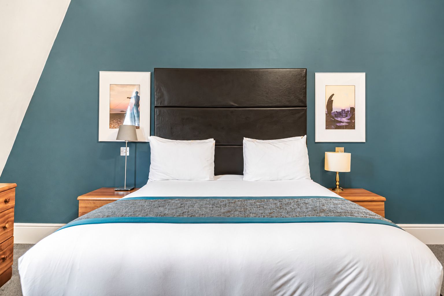 Ein Hotelzimmer mit einem großen Bett, einem dunklen Kopfteil, zwei Nachttischen mit Lampen und zwei gerahmten Bildern an den Wänden.