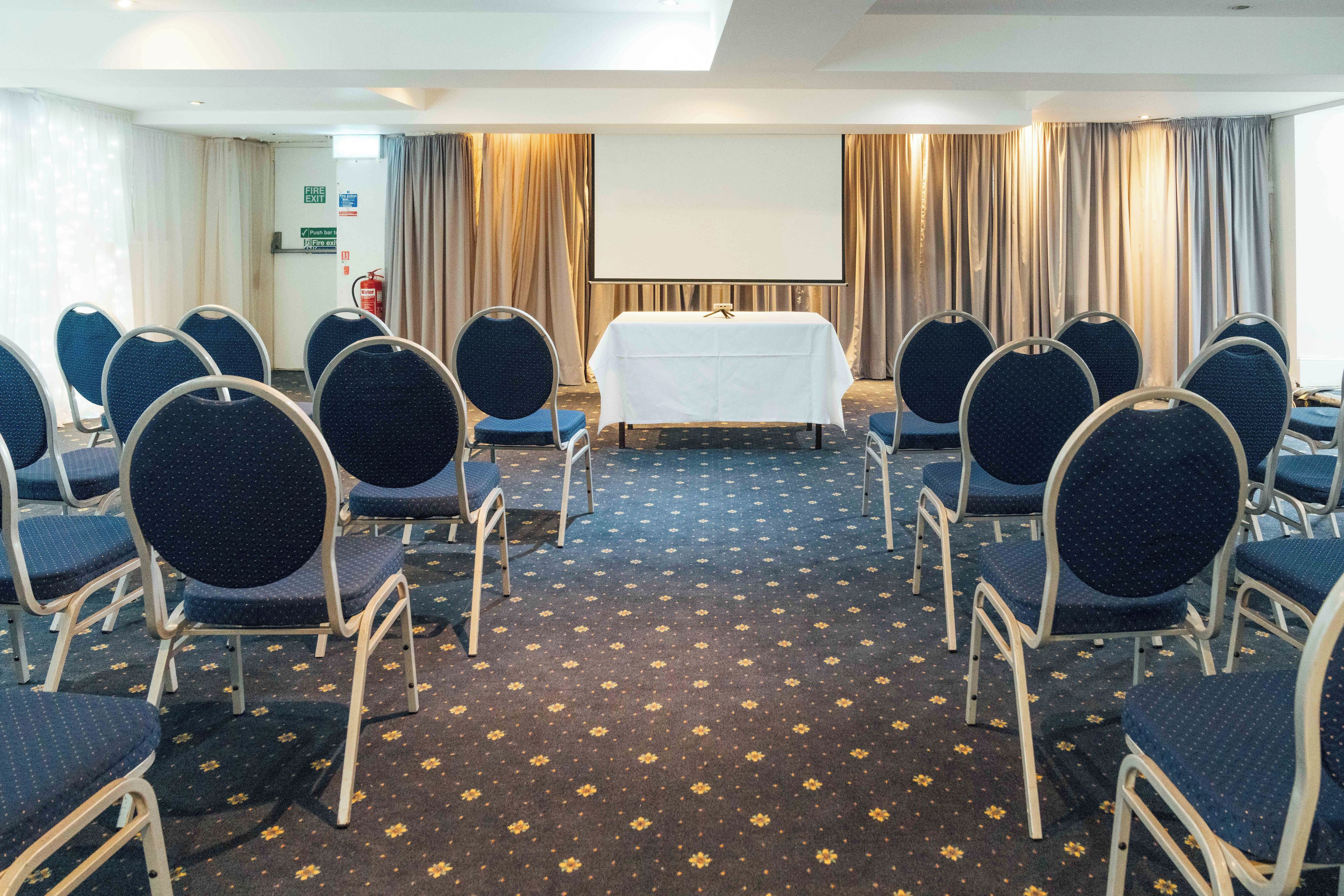 Une salle de conférence avec des rangées de chaises disposées face à une table sur laquelle se trouve un écran de projection, un panneau indiquant la sortie de secours et des rideaux.