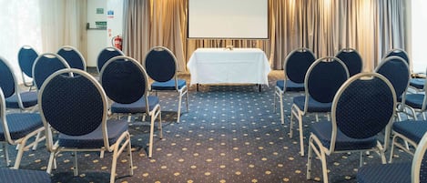 Une salle de conférence avec des rangées de chaises disposées face à une table sur laquelle se trouve un écran de projection, un panneau indiquant la sortie de secours et des rideaux.