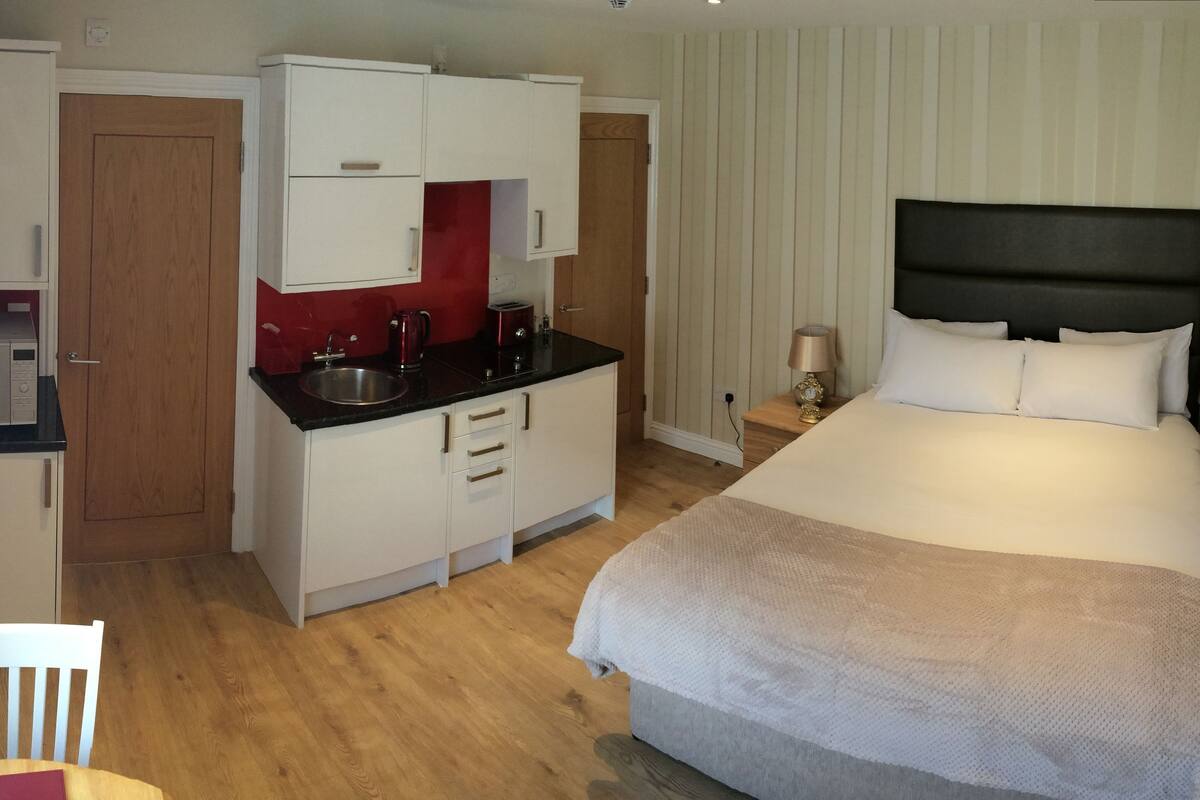 A compact hotel room with a bed, a small kitchenette, a wooden chair, and a bedside table with a lamp.