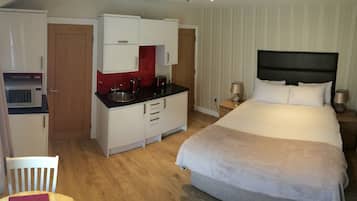 A compact hotel room with a bed, a small kitchenette, a wooden chair, and a bedside table with a lamp.
