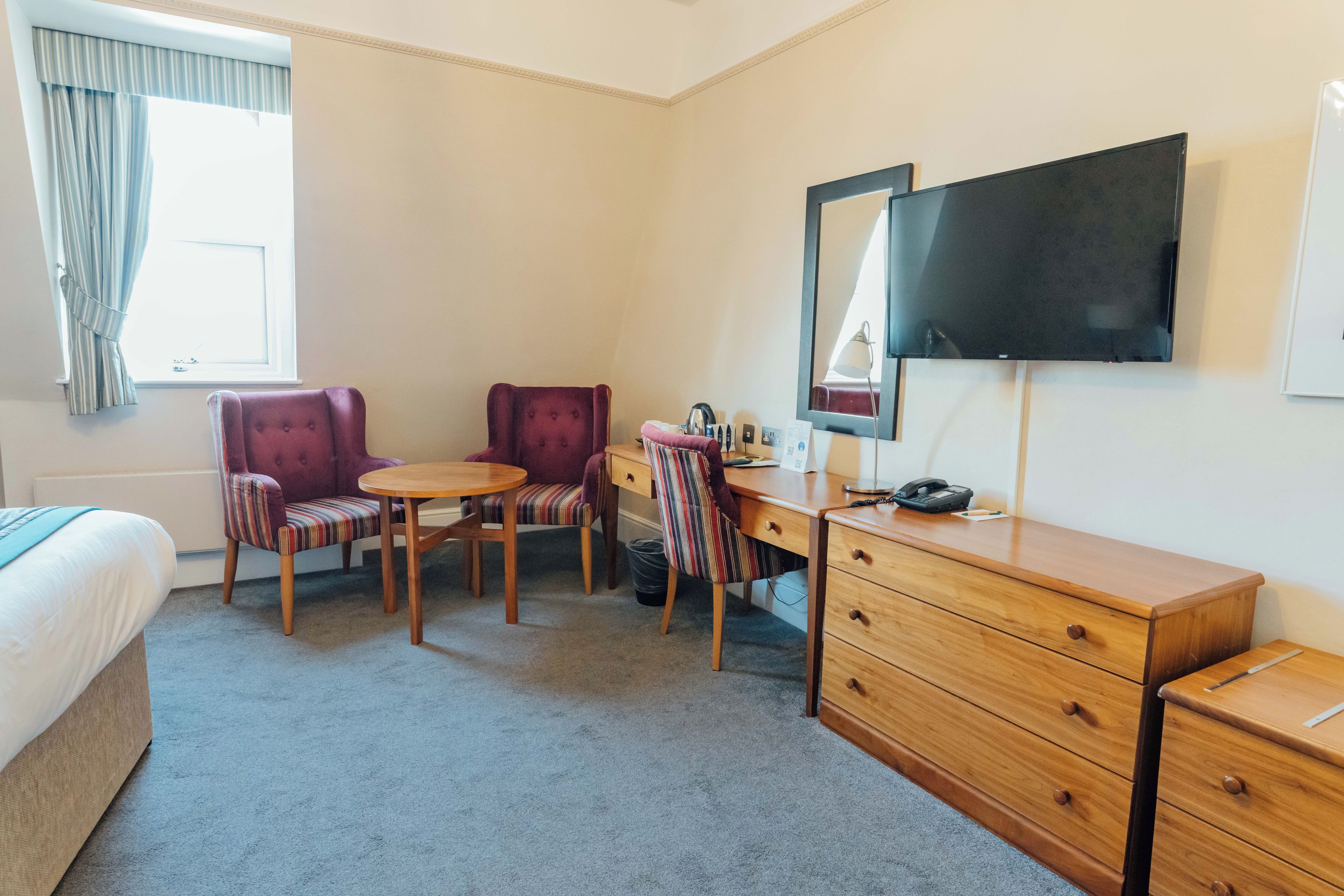 Ett hotellrum med en säng, två fåtöljer, ett litet runt bord, en byrå i trä med lådor, en platt-TV som är monterad på väggen och ett fönster med gardiner.