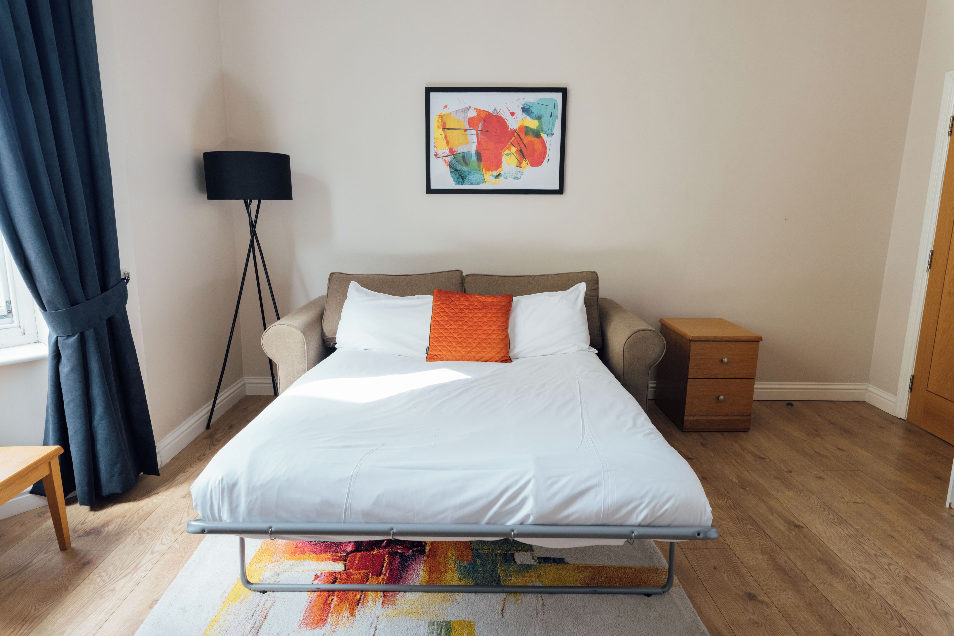 Un dormitorio ordenado con una cama, mesita de noche, lámpara y una alfombra colorida.