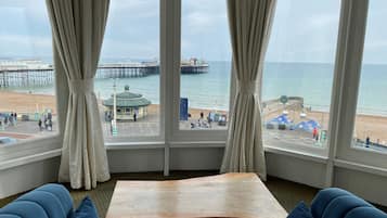 Une pièce dotée d’une grande fenêtre donnant sur une plage, d’une table en bois et de sièges recouverts d’un tissu bleu.