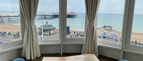 A room with a large window overlooking a beach, a wooden table, and blue upholstered seating.