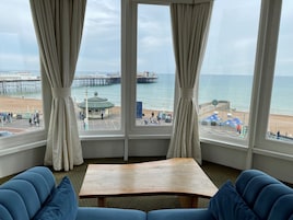 A room with a large window overlooking a beach, a wooden table, and blue upholstered seating.