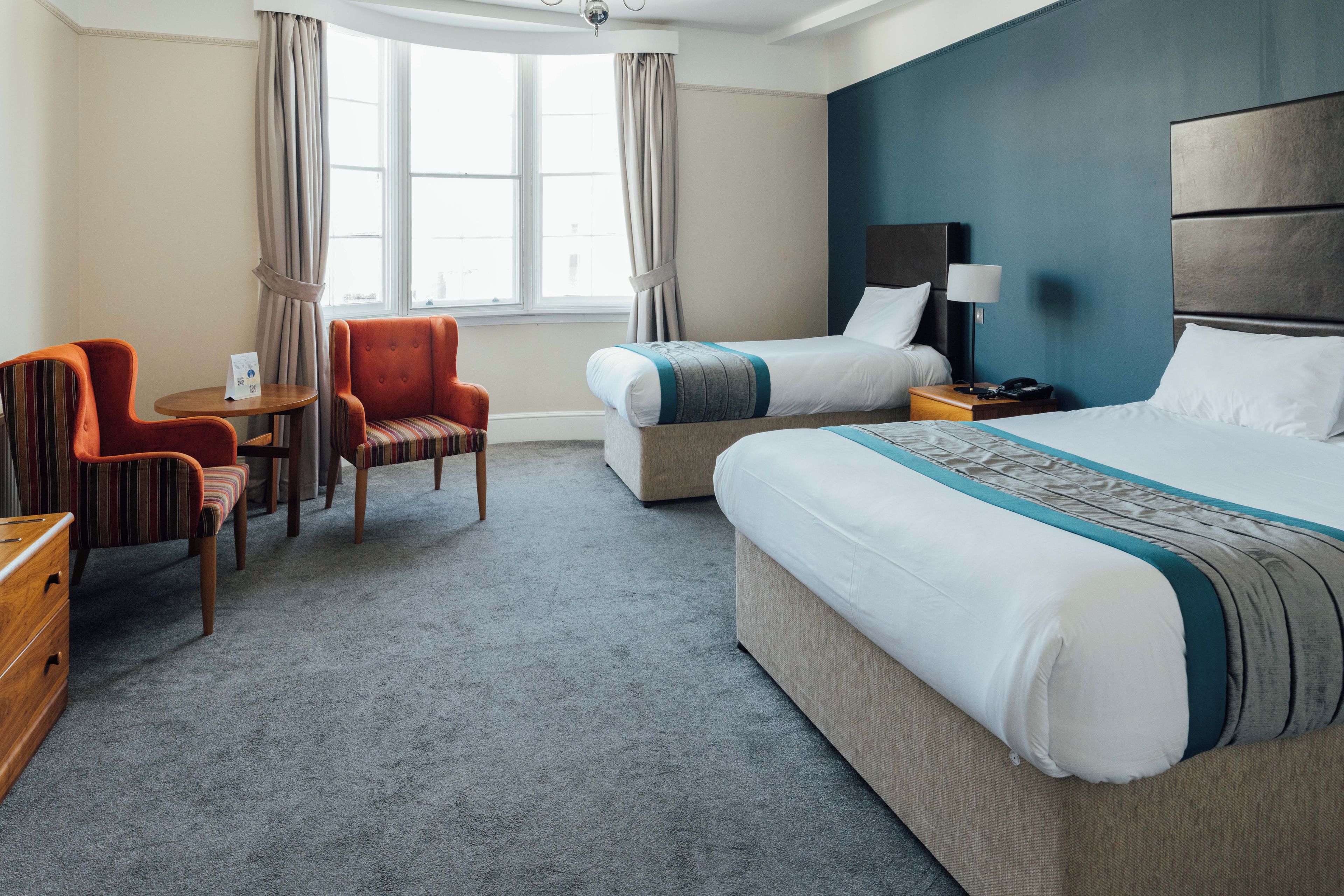 Una habitación de hotel con una cama grande, otra más pequeña, un escritorio, dos sillas, una lámpara y un ventanal con cortinas.