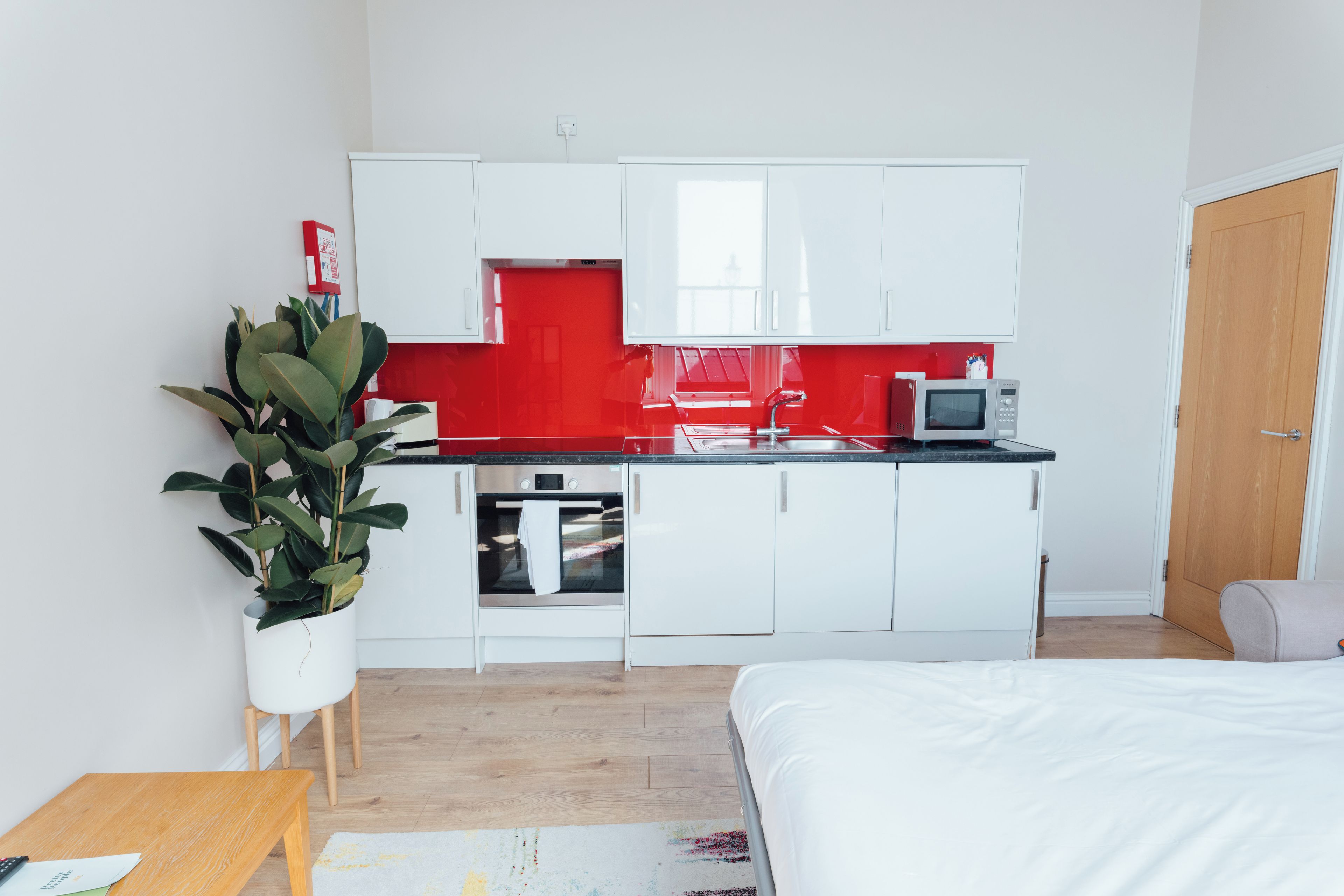 A compact kitchenette with white cabinets, a microwave, and a small oven. There is a potted plant and a wooden table with a chair.