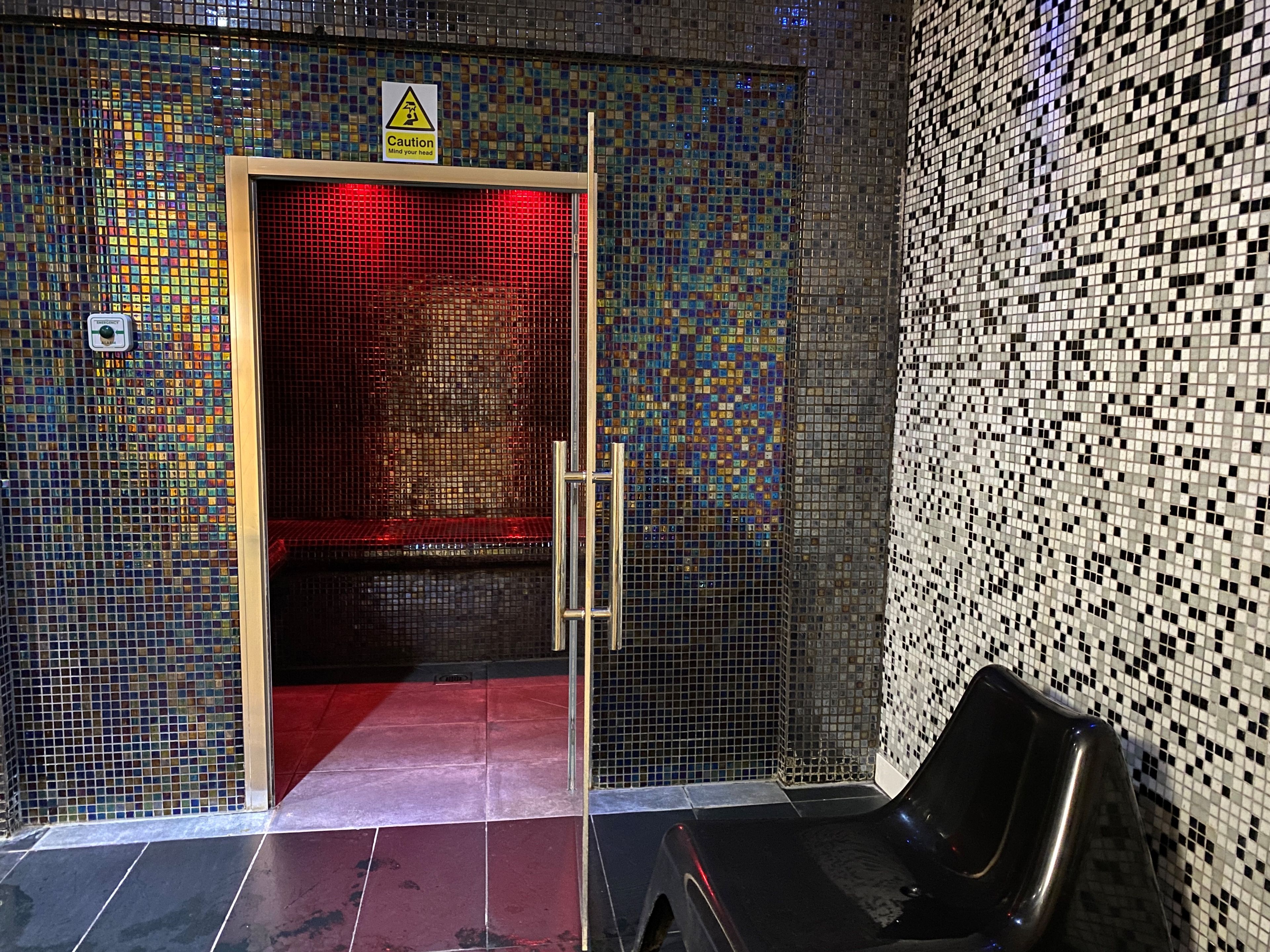 A tiled wall with a caution sign, a door with a red frame, and a black object on the floor.