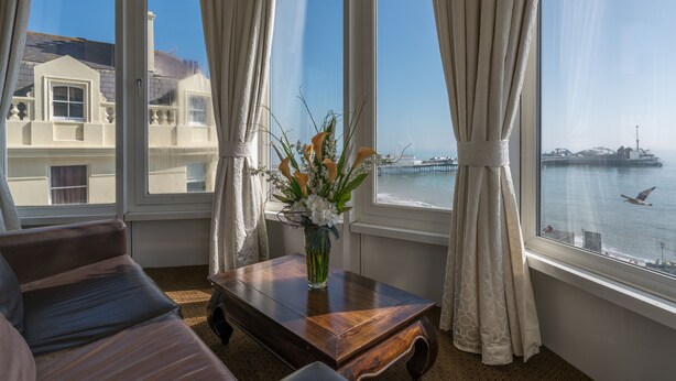 A room with a large window, a wooden table with a vase of flowers, and a view of a body of water and buildings. - Queens Hotel (Brighton)