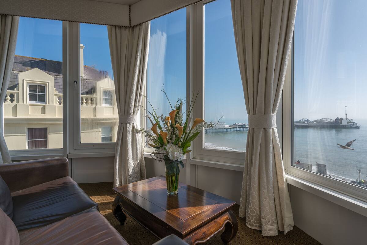 A room with a large window, a wooden table with a vase of flowers, and a view of a body of water and buildings.