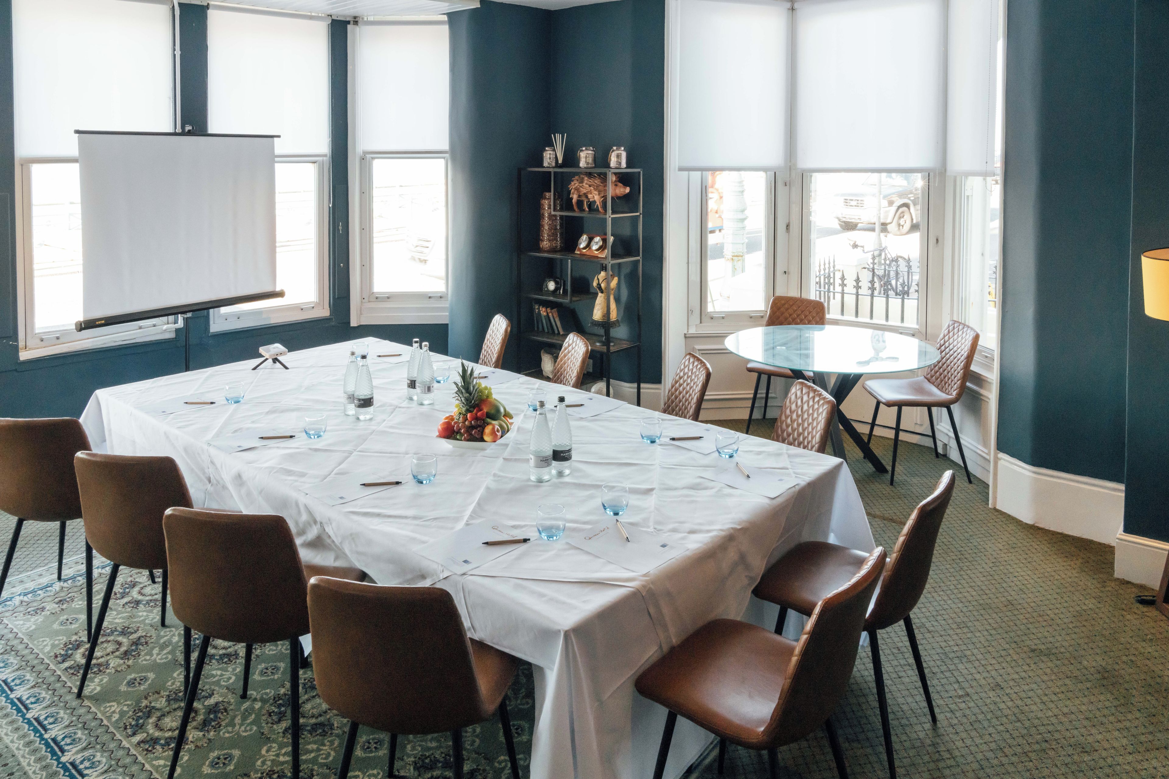 A conference room with a long table, chairs, a projector screen, and a bookshelf.