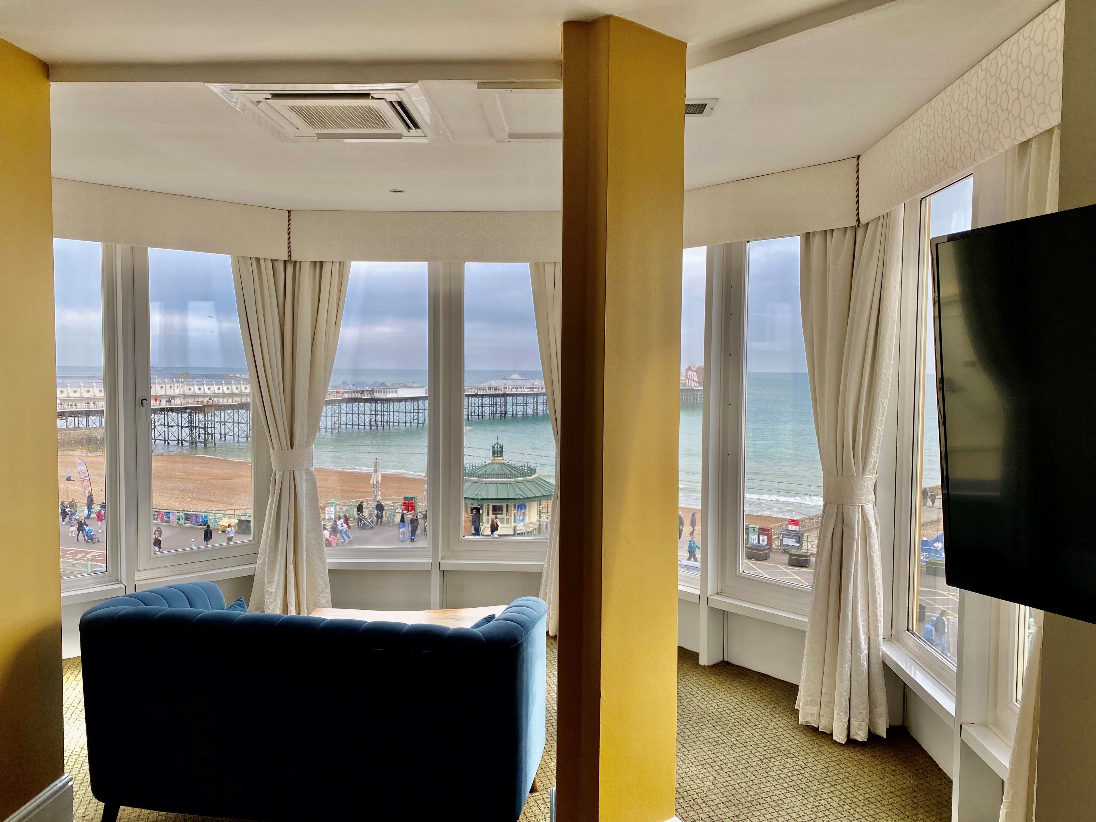 A hotel room with a large window offering a view of a pier and beach, a flat-screen TV, and a blue sofa.
