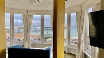 A hotel room with a large window offering a view of a pier and beach, a flat-screen TV, and a blue sofa.