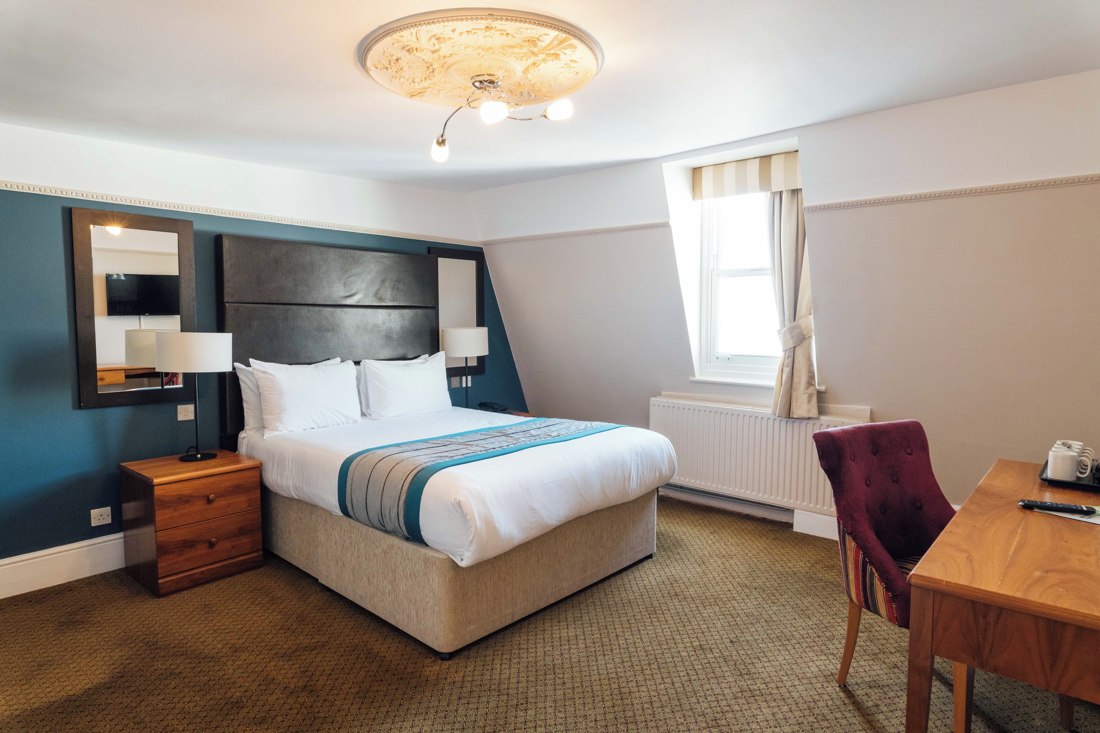 A hotel room with a bed, bedside tables, a desk, a chair, a television, and a window with curtains.