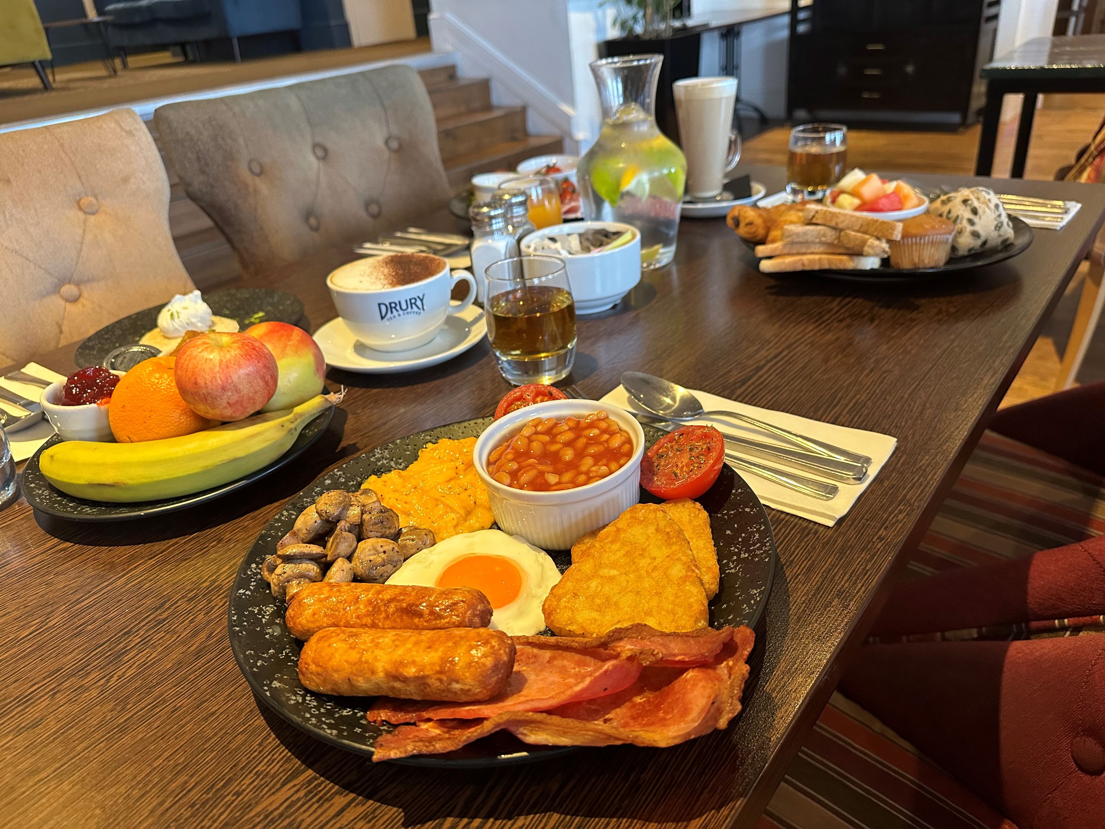 A breakfast table with plates of eggs, bacon, beans, toast, and fruit.