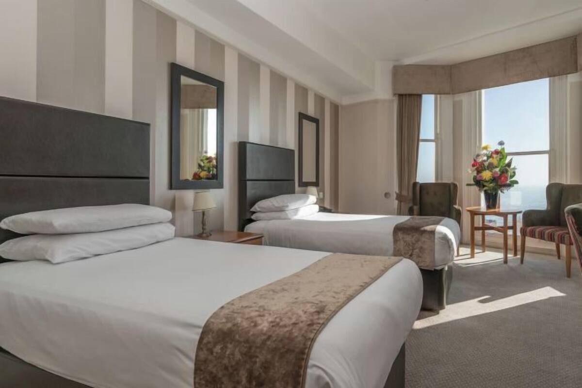 A hotel room with two beds, a TV, a chair, a table, and a view of the sea.