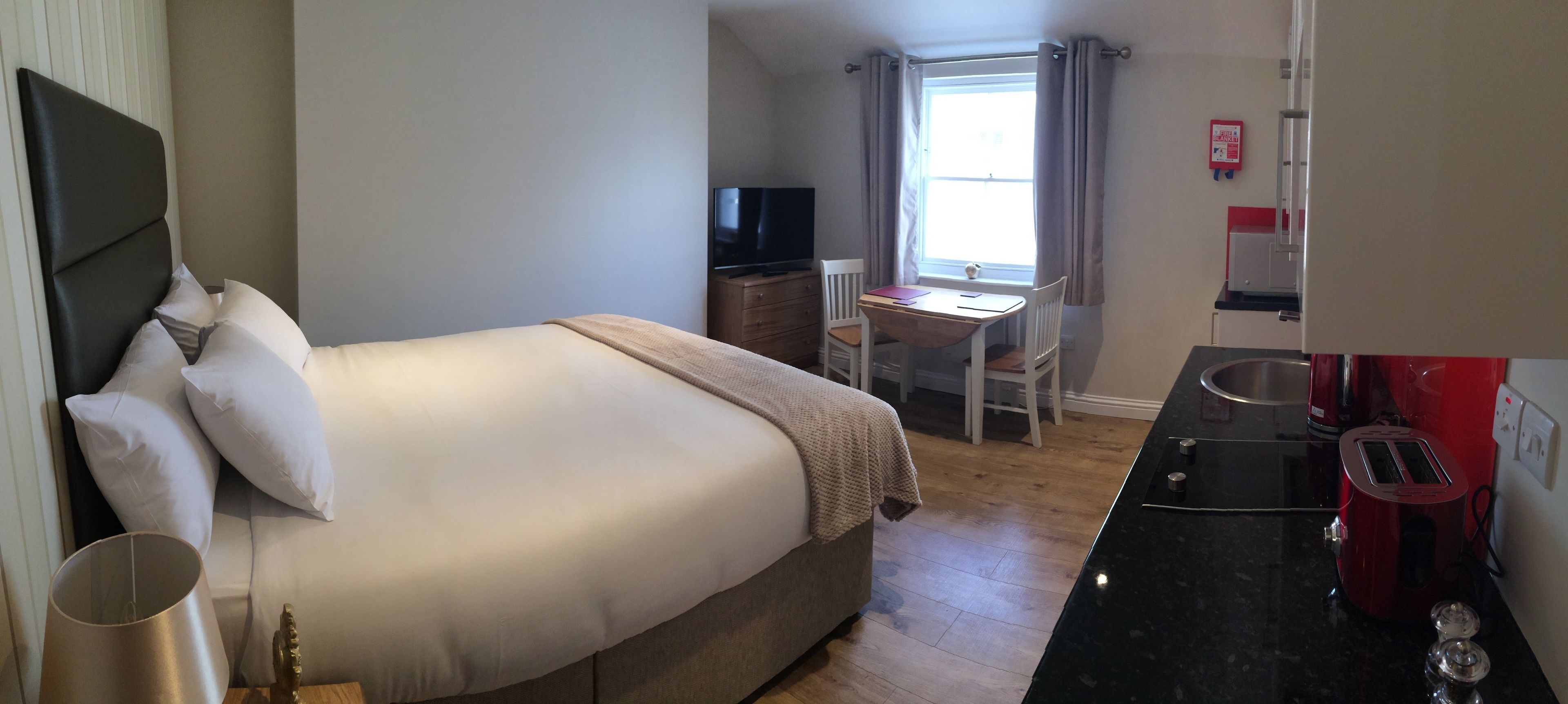 A bedroom with a bed, bedside table, lamp, and a small desk with a chair. There is a window with curtains, a television, and a red toaster on the counter.